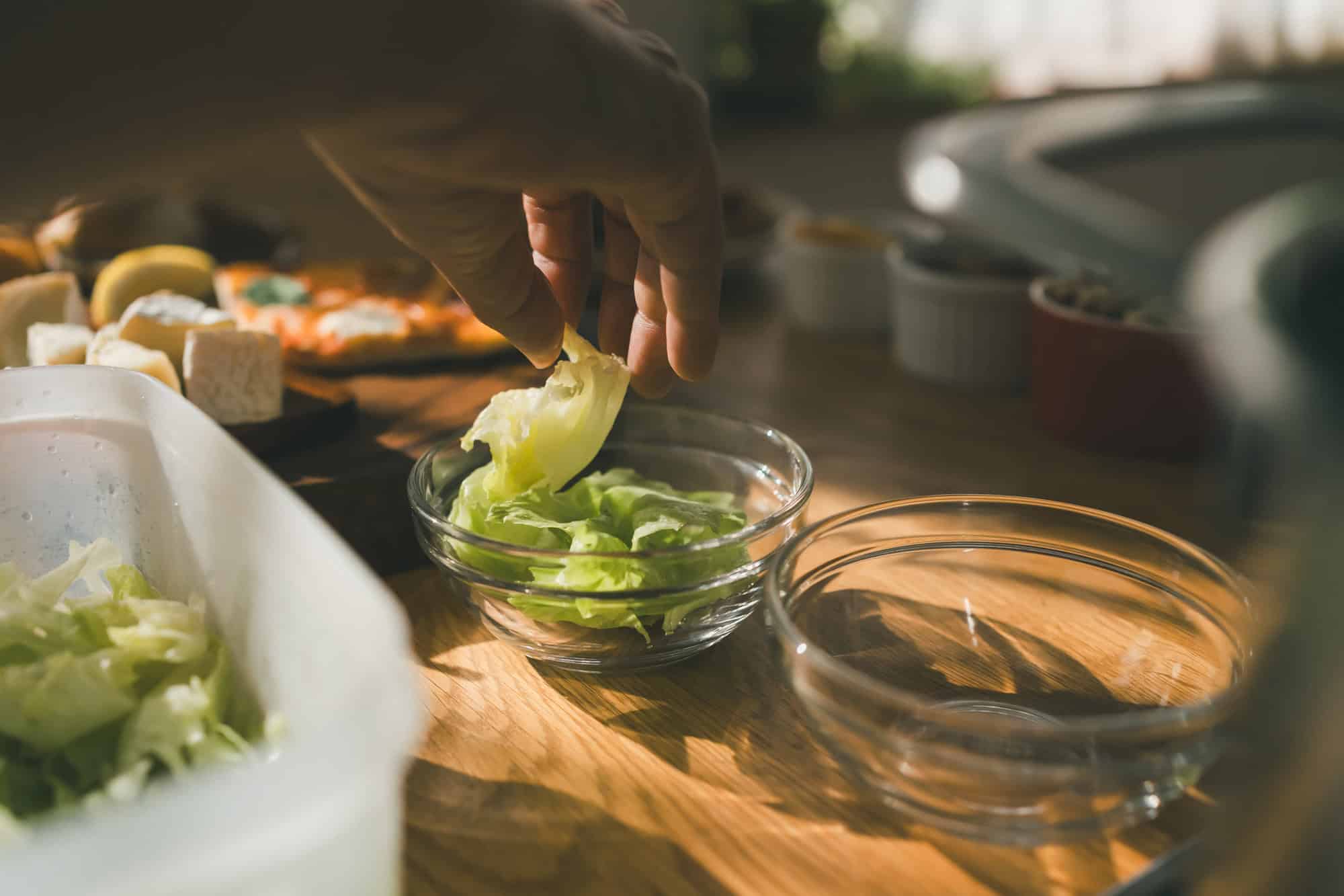 A wooden table set with plates of assorted salads, a bowl of soup, slices of bread on a wooden board, and a glass carafe filled with water, lemon, and mint, with a blurred garden visible in the background.