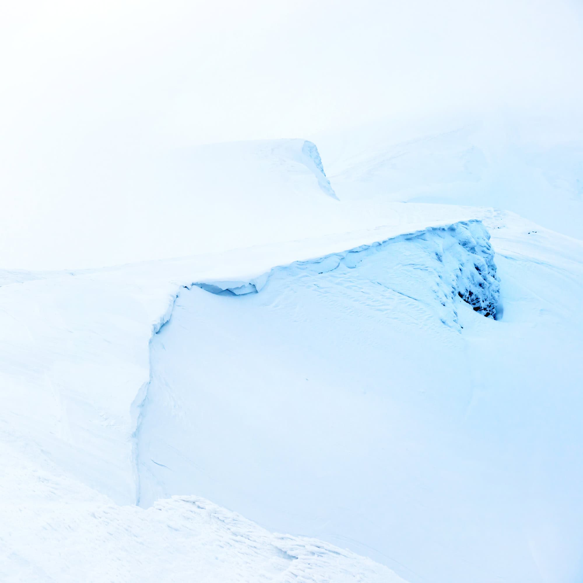 A snowy, windswept landscape with a dramatic ice cliff and deep crevasse. The scene is bright, dominated by white snow and blue shadows, evoking a cold, remote, and rugged Arctic or Antarctic environment.