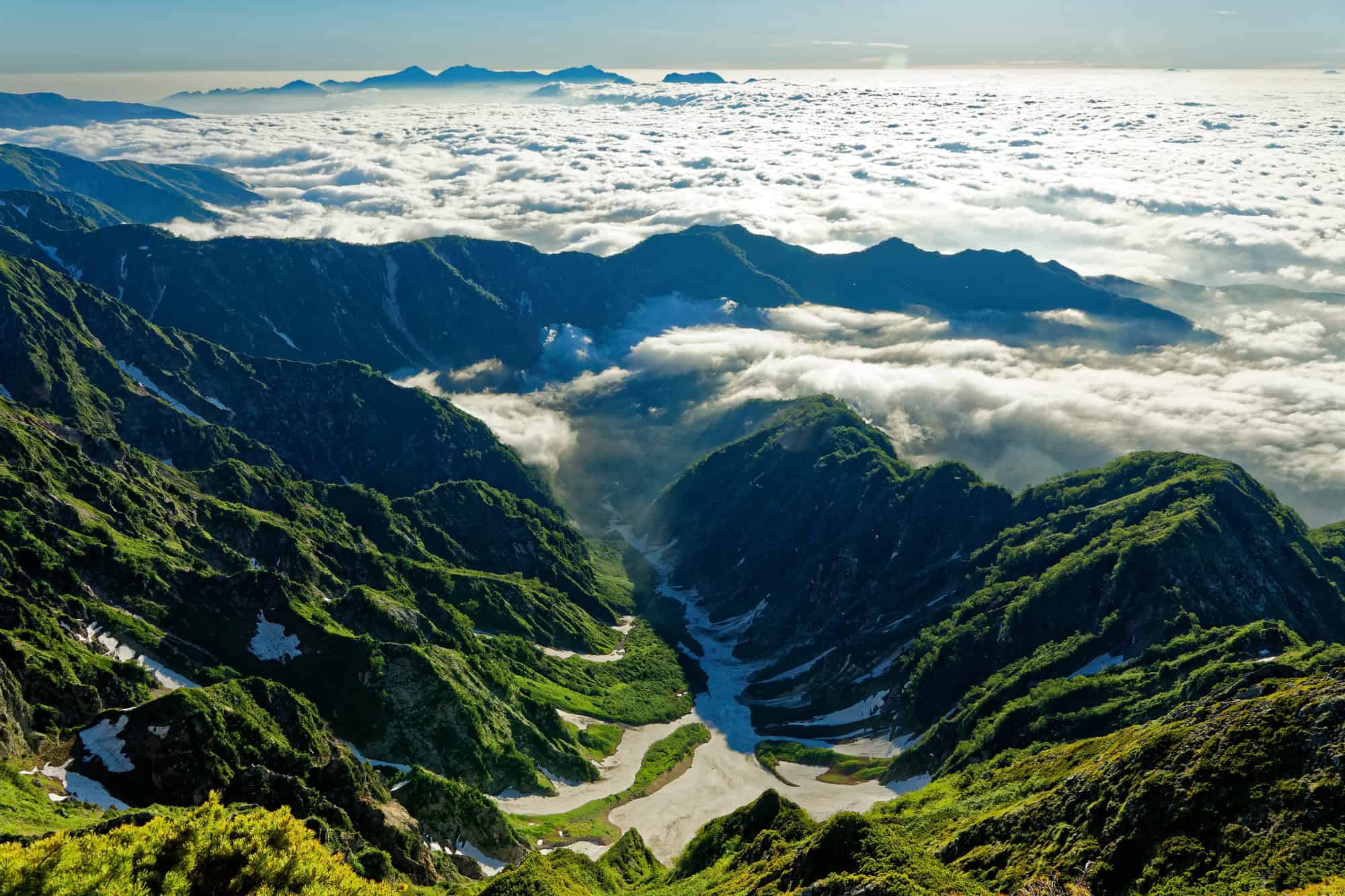 A lush green mountain valley with patches of snow, overlooking a sea of clouds filling the lower landscape, with distant mountain peaks visible on the horizon under a clear sky.